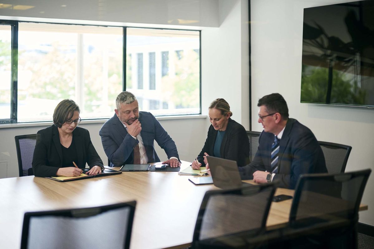 a group of four people sitting around a boardroom table in an office looking at documents on the table