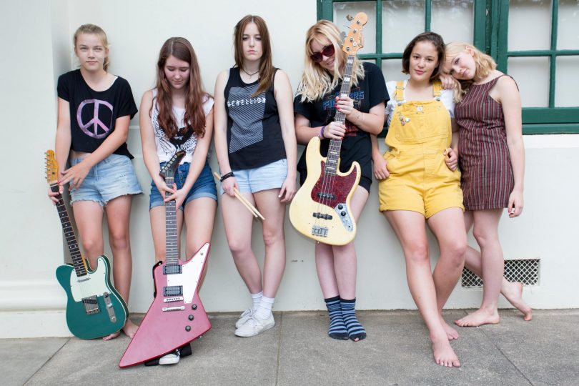 Photograph of six members of a band in front of a wall holding guitars