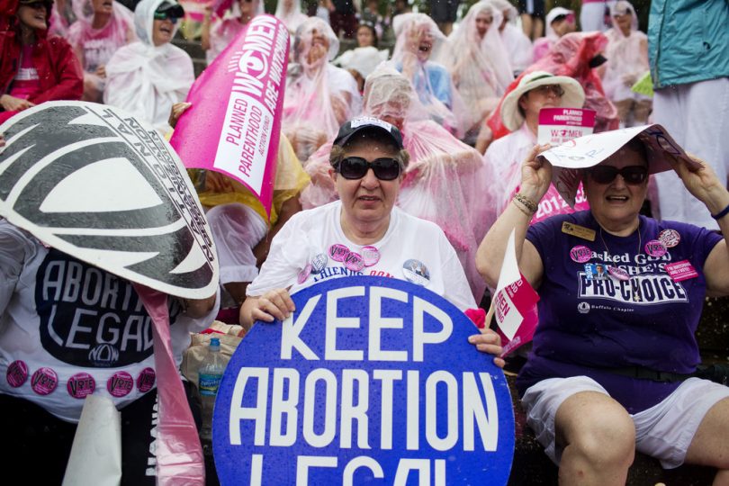 Two middle aged women sitting in front of a group at a protest with a sign that reads "Keep Abortion Legal"