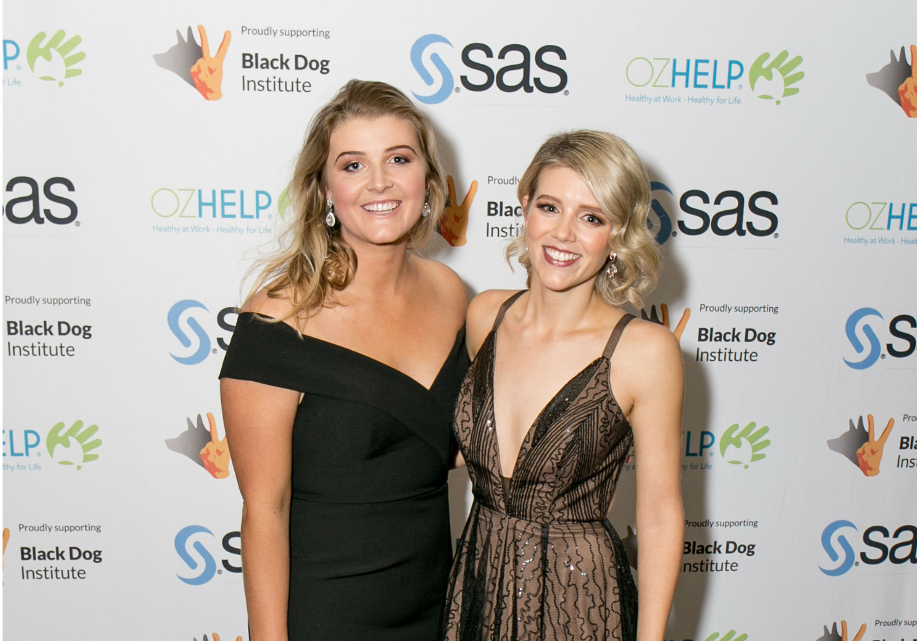 Britt Shephard and Shannon Narracott in front of sponsors logos at the Suicide Awareness Ball, smiling at camera