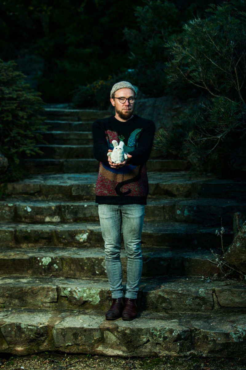 Canberra musician, Sebasian Field, stands in a dark outdoors setting, at the end of old stone steps. He holds a small, white, toy rabbit and wears jeans, a dark knitted jumper and light beanie.