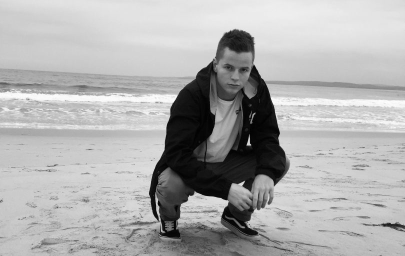 Photograph shows rapper Cappo crouching on a beach, black and white