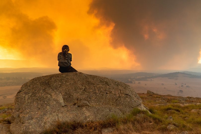 Girl sitting on rock watching bushfire in distance.
