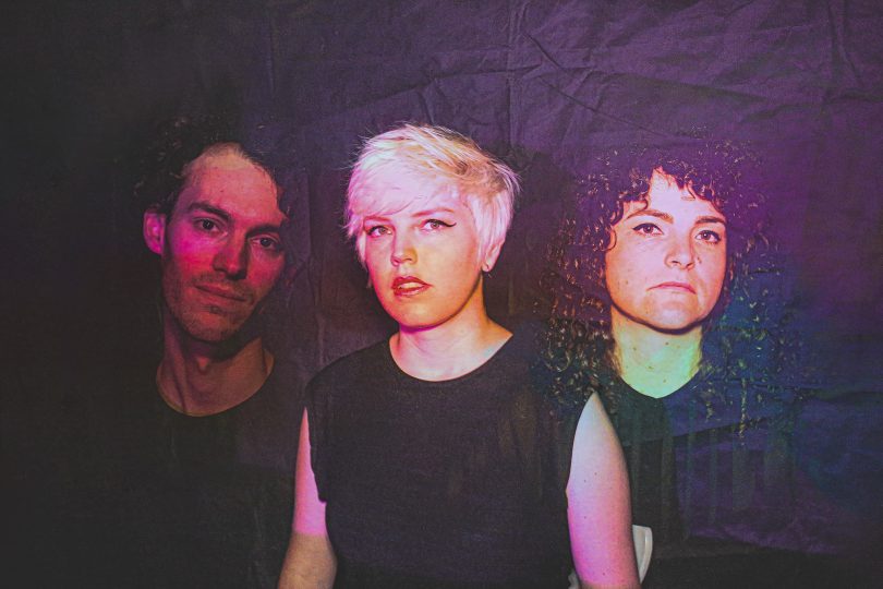 Photograph shows three members of Cable Ties looking at camera on purple backdrop