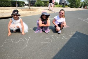 Three children kneeling on street in front of "Play Street" written in chalk.