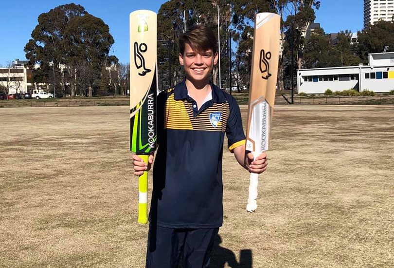 Young boy standing on oval holding two cricket bats.