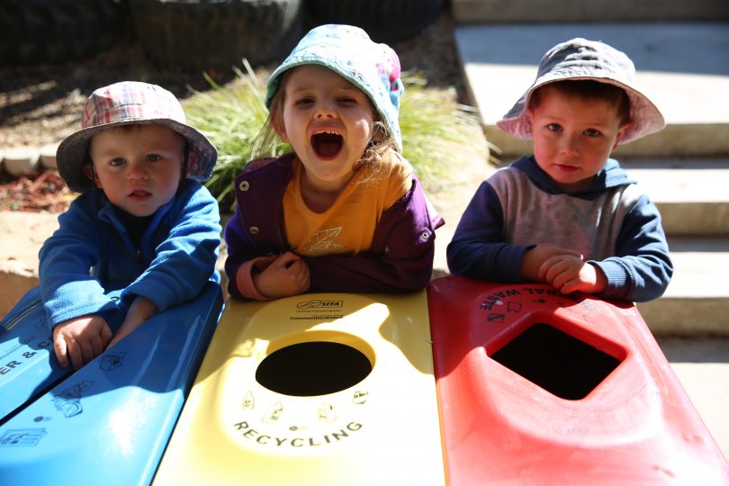 Three children leaning on recycling bins.