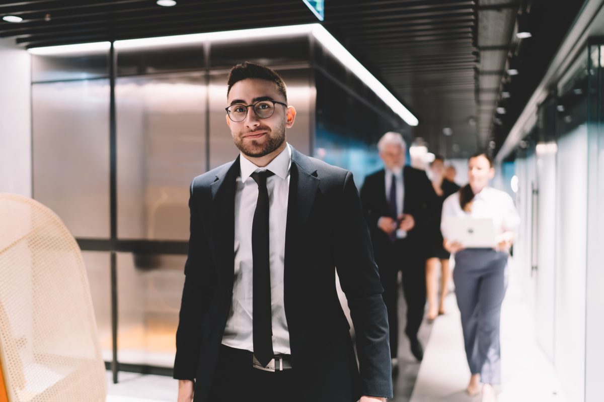 Smiling young male entrepreneur in formal wear and eyeglasses walking in corridor of modern office and looking at camera while waiting for colleagues