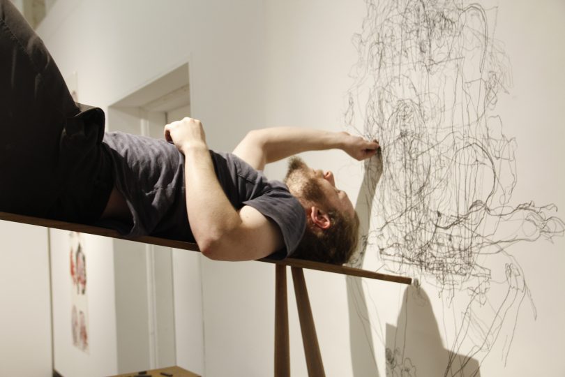 Artist Robbie Karmel lying on a bench on his back creating an abstract drawing with charcoal on wall.