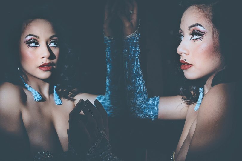 A woman with blue velvet gloves and blue tassel earrings leans against a mirror in a darkened space