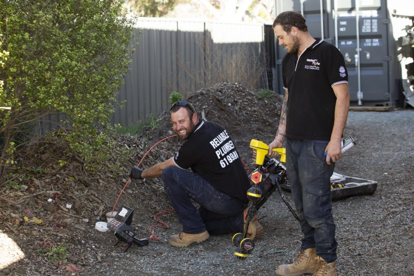 one man crouching and another man standing, both holding tools and working on pipes underground