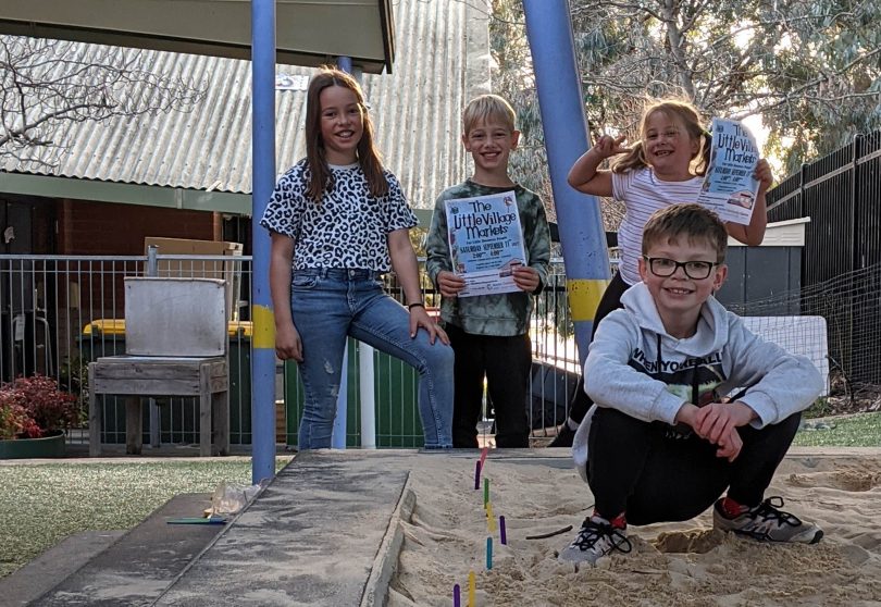 Lucy Swift with her siblings and friends in a sand pit holding flyer for The Little Village Markets