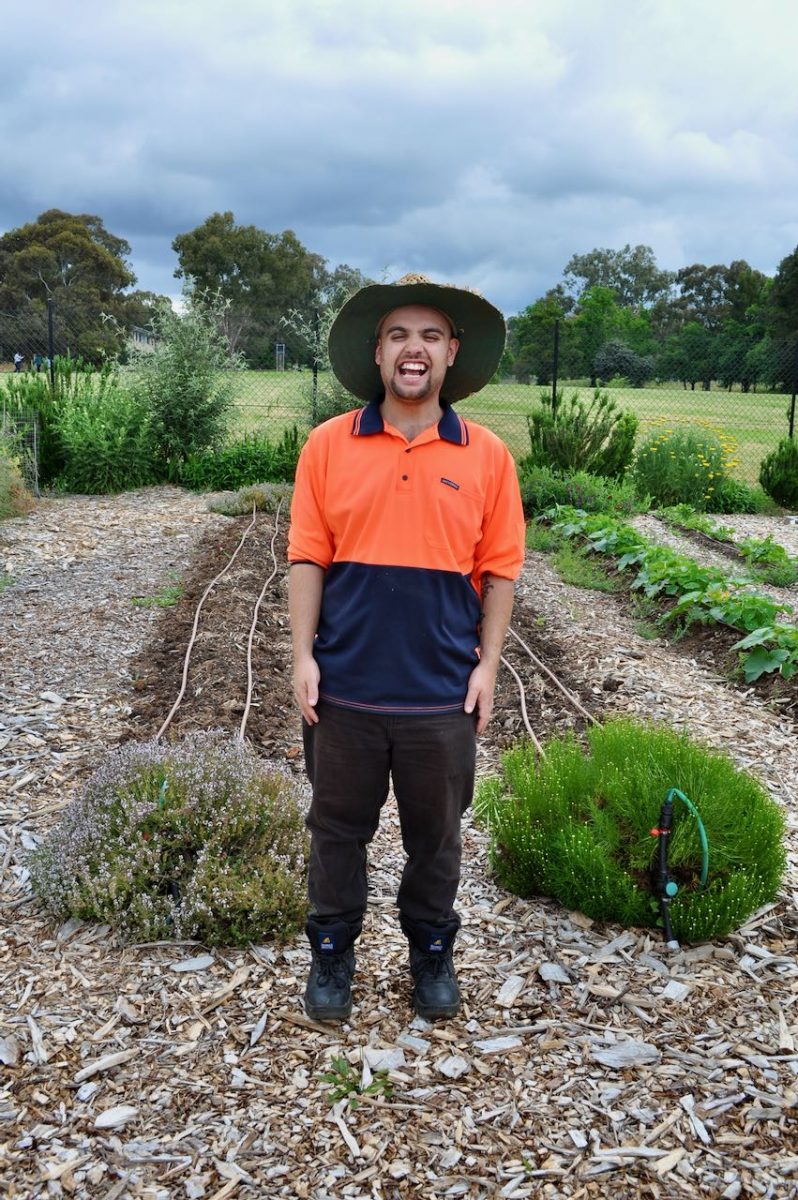 Young man in high vis smiles broadly in front of garden beds