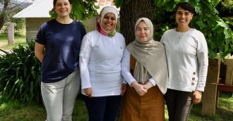 Four smiling women, two wearing headscarves, stand under a tree