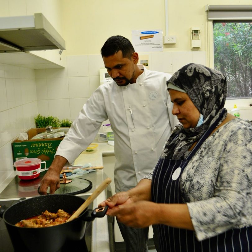 A chef and a woman wearing an apron and hijab stir a pot on the stove