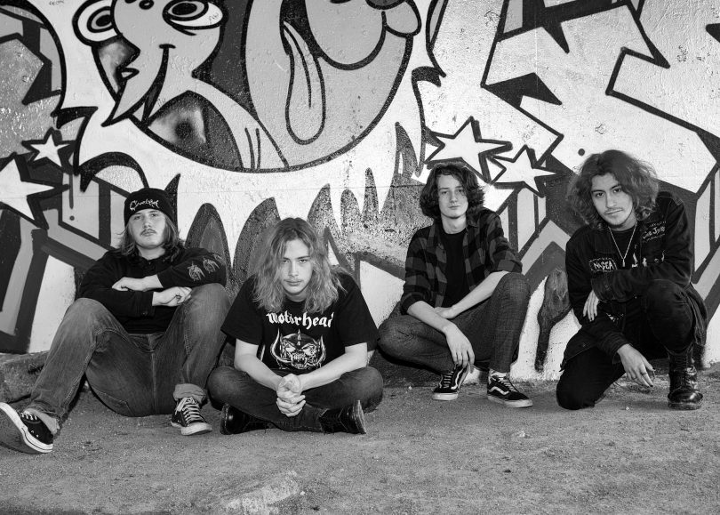 Image shows four youths sitting in front of street art wall. Black and white.