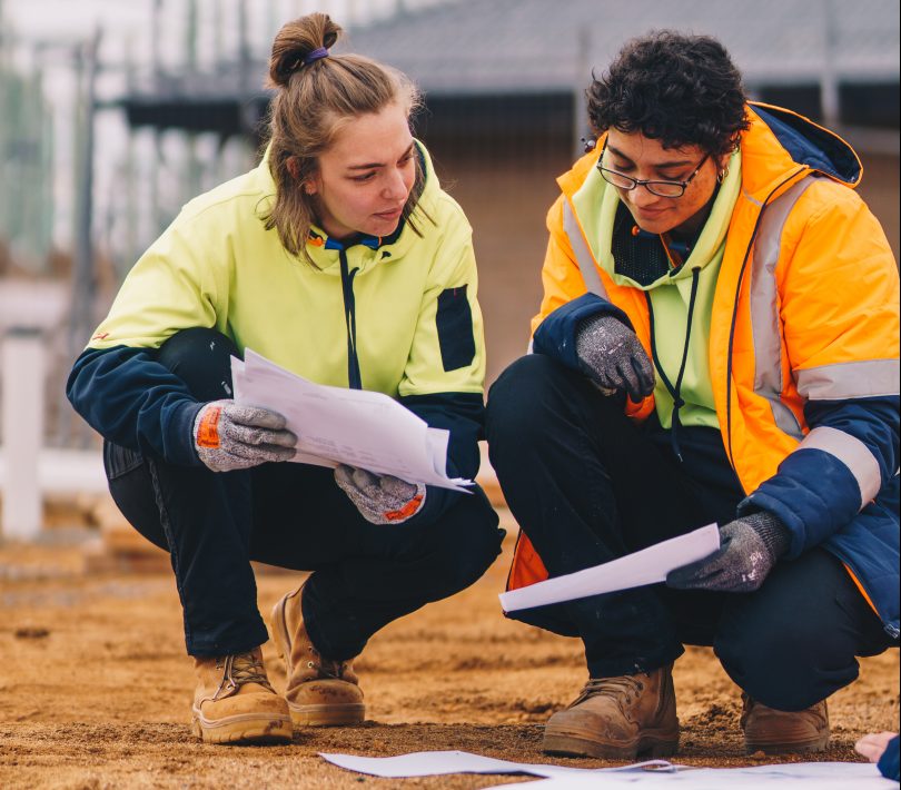 Two women in high visibility vests crouched in a construction site