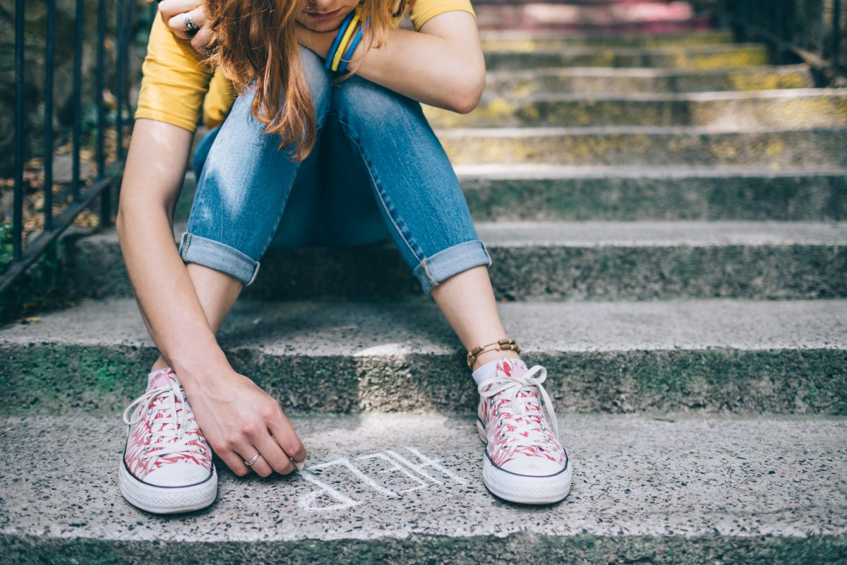 Depressed looking teenage girl sitting on steps, writing 'HELP' in chalk on the bottom step