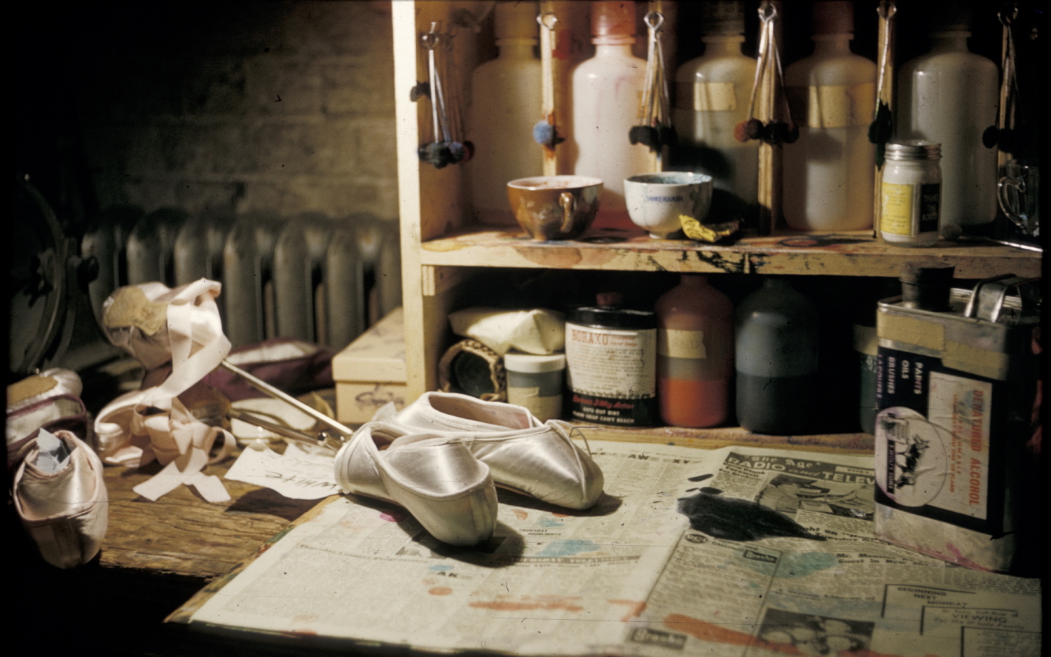 Ballet slippers sit on a workshop table in front of a cupboard with craft materials.