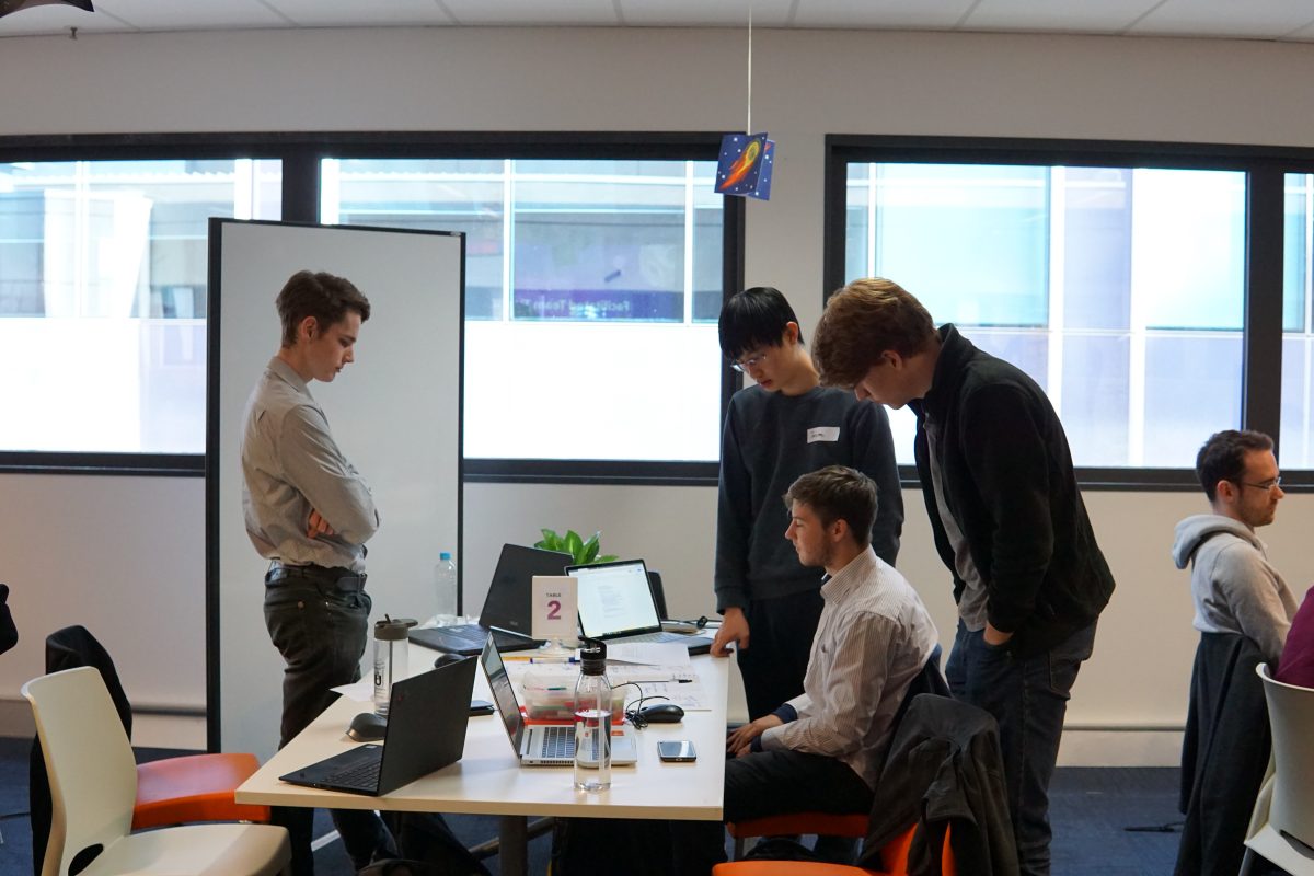 Four young men peer at two open laptops on a table.