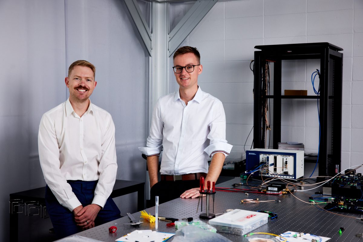 Vai Photonics' two founders stand behind a table filled with electronics.