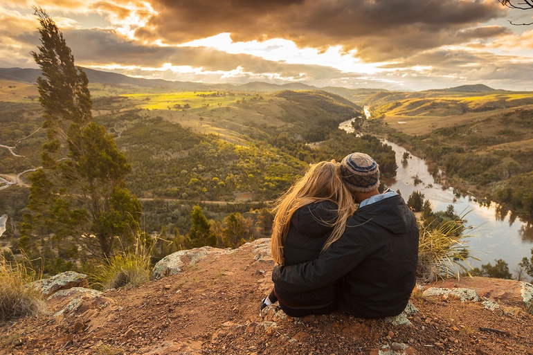 A couple sitting arm in arm overlook a river and rolling hills.