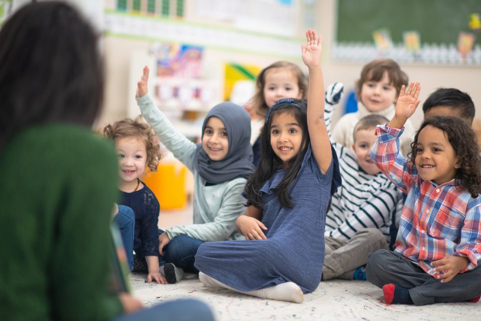 A multi-ethnic group of preschool kids is indoors in their classroom. They are sitting on the floor and raising their hand to answer a question posed by their teacher while reading a story