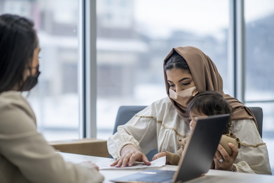  A young Muslim woman, with her young daughter sitting on her lap, speaks to an office worker