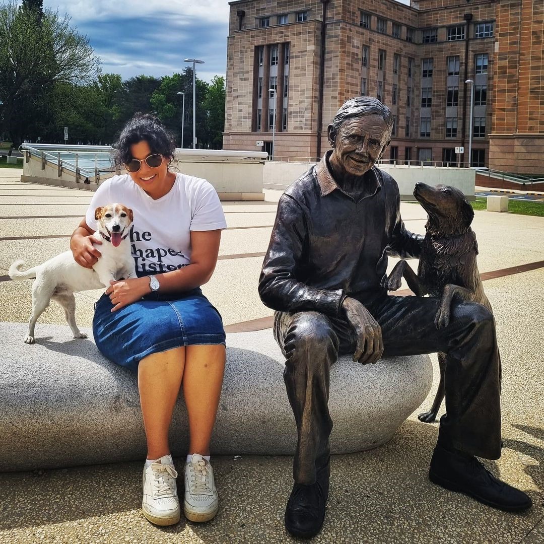 Woman with dog sitttting next to statue of John Gorton with his dog. 