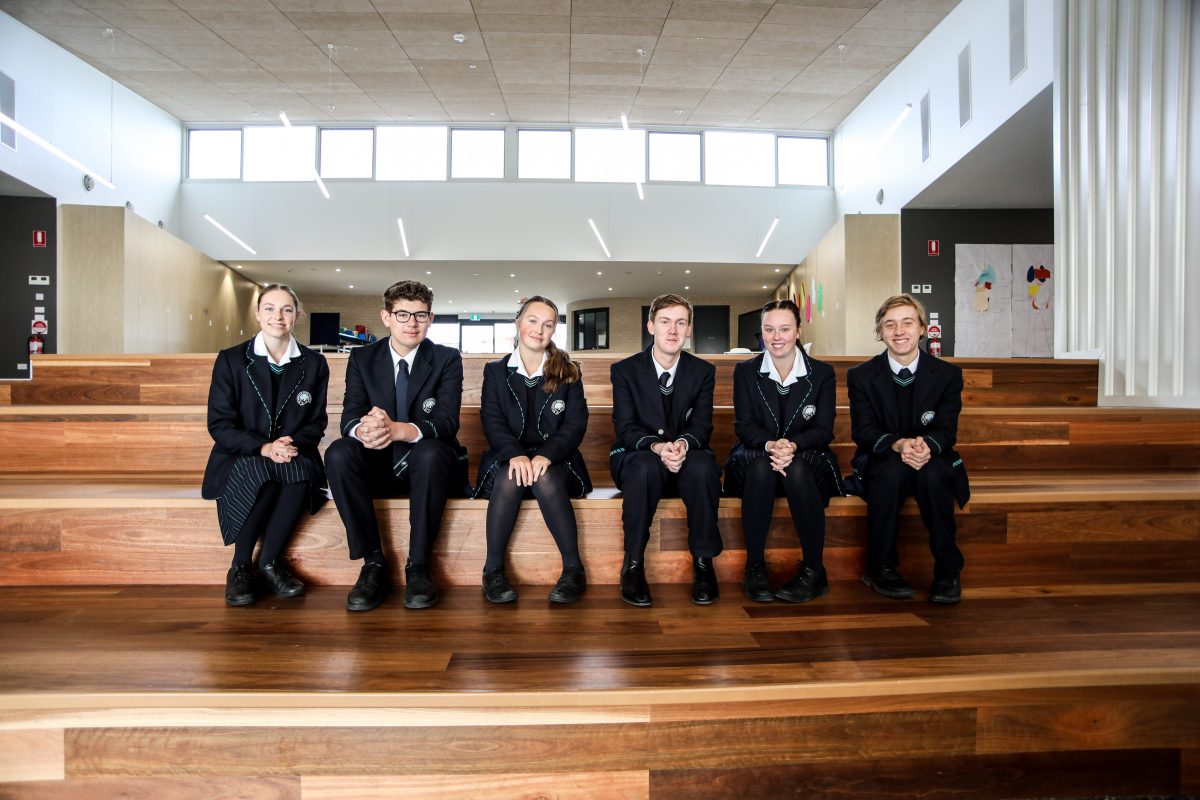 Six students sit side by side on wooden stairs inside a large room.