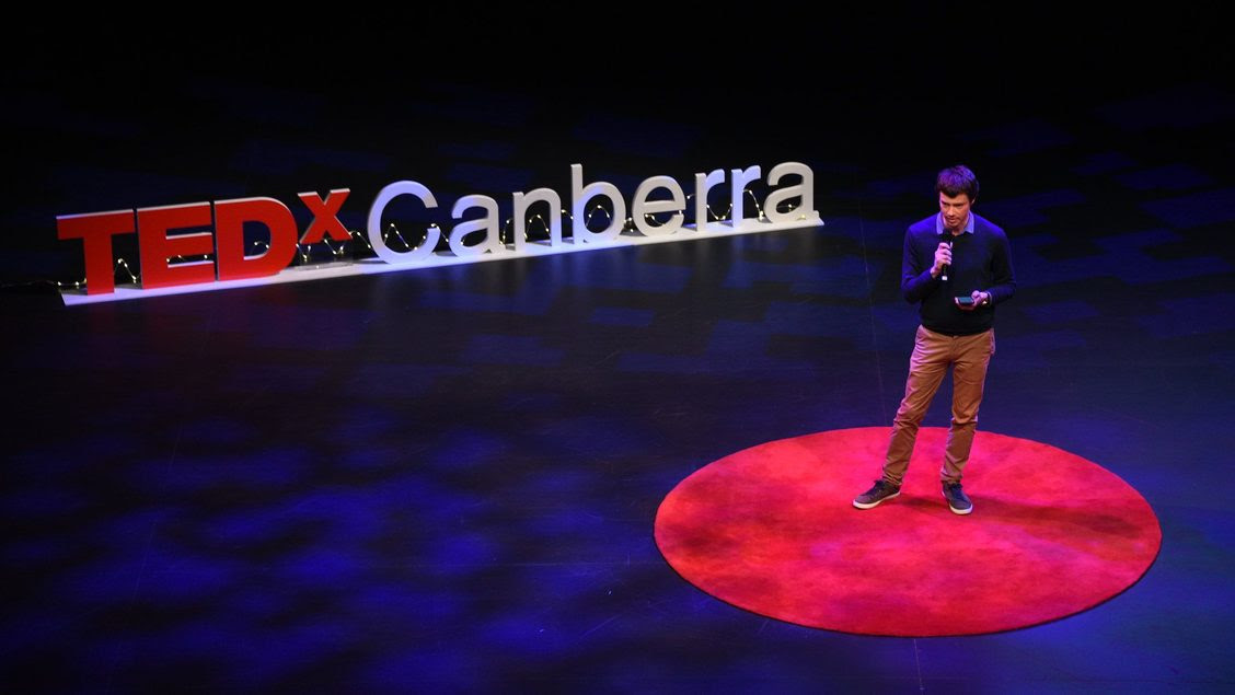 Man speaking into a microphone on stage next to a TEDxCanberra sign