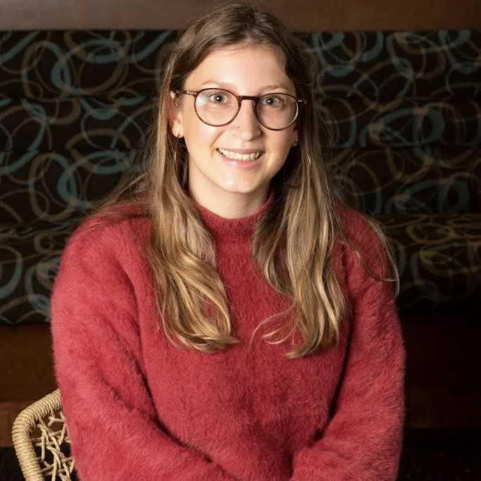 A smiling woman with round glasses wearing a red jumper