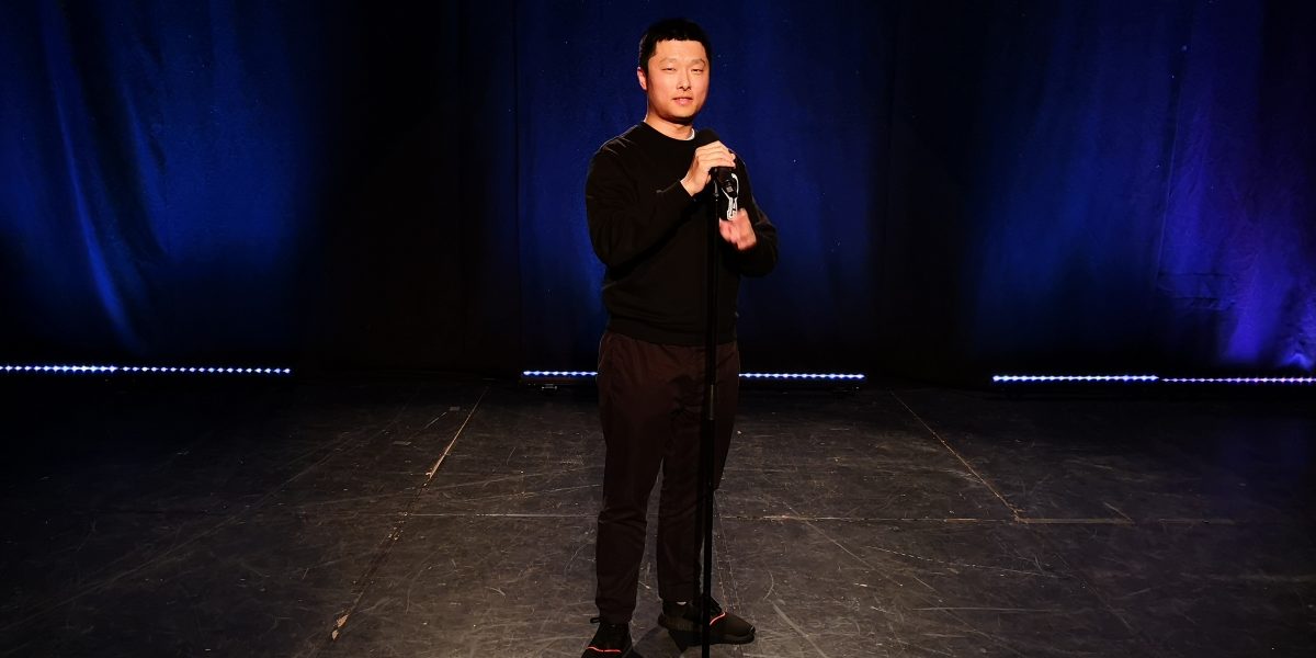 Comedian Jeff Shen on a stage with a blue curtain behind him.