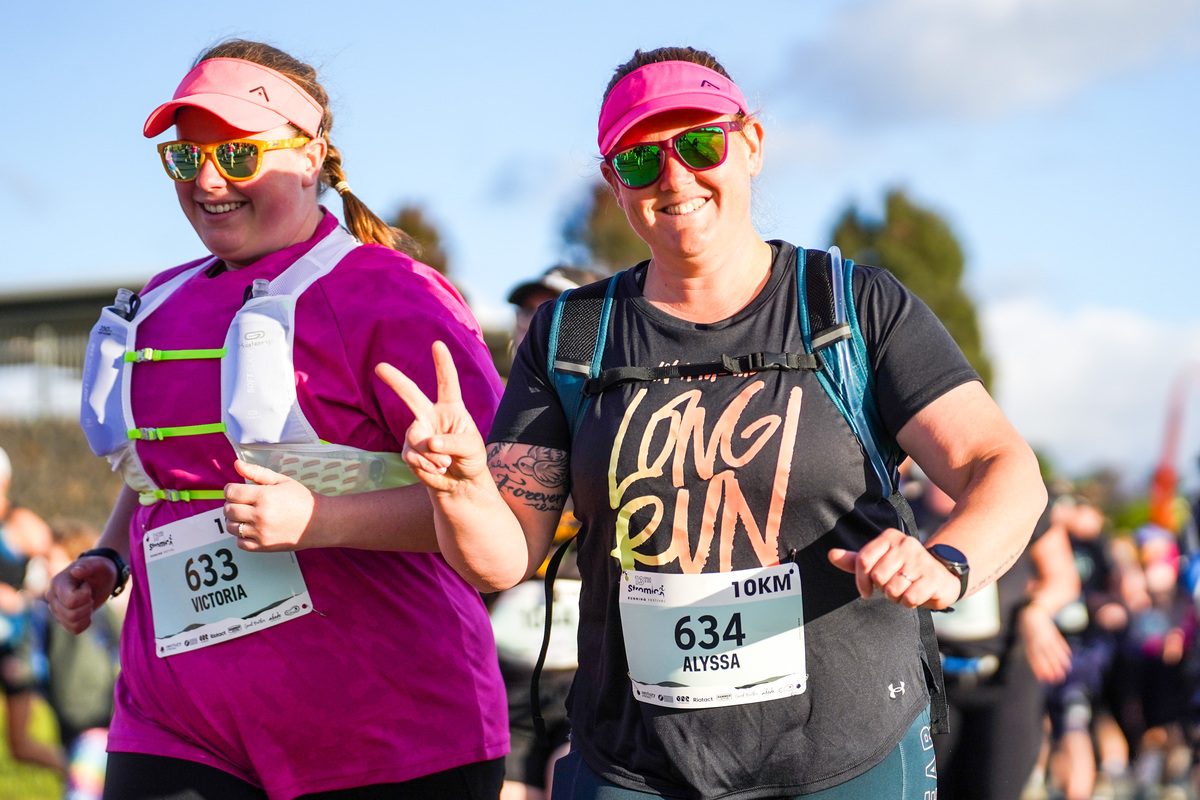 Two runners running past, with one in the front holding up their right hand with the peace sign.