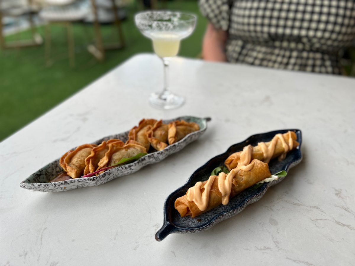 Dish of spring rolls covered with sauce, dish of fried dumplings and cocktail in background on outdoor terrace at Yaki Boi
