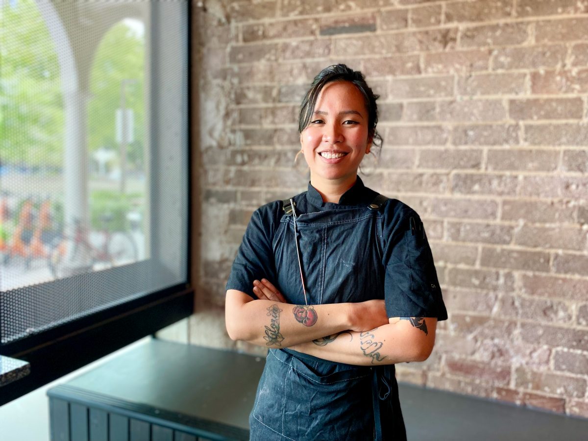 Chef Adi wears black chef's jacket and apron and smiles with arms crossed against a brick wall at Luna