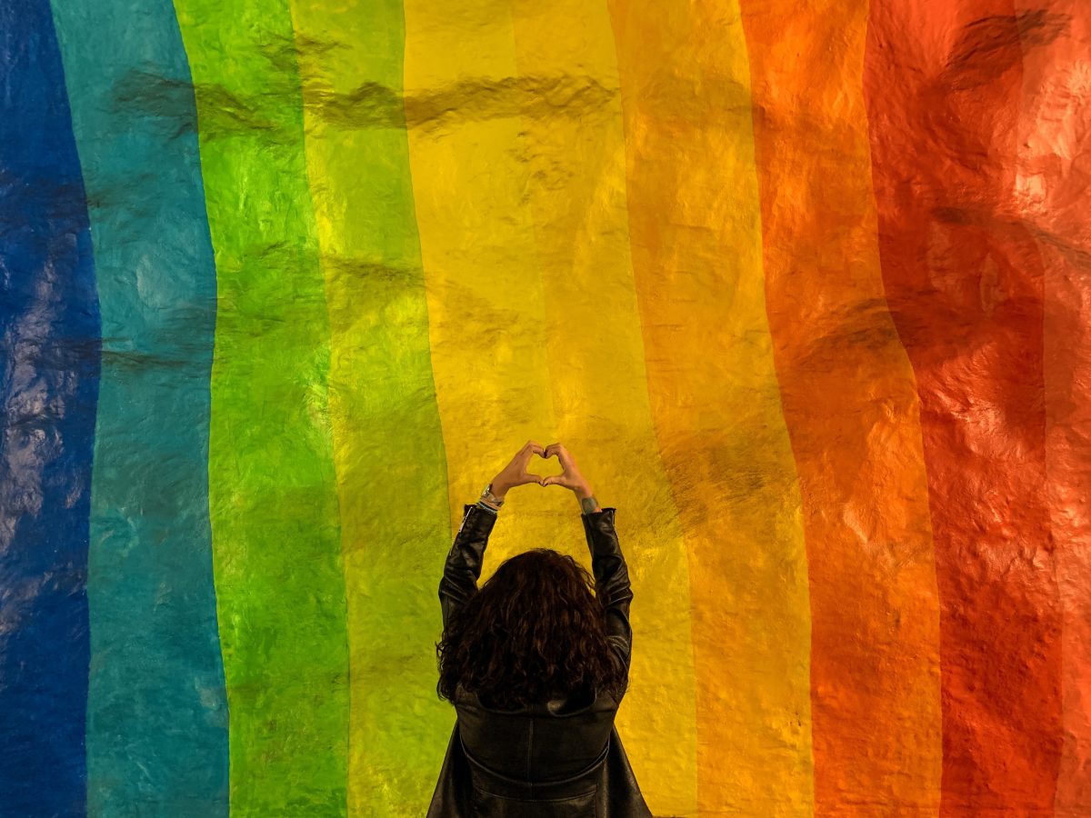 Person from behind forming a heart shape with their hands in front of a rainbow backdrop