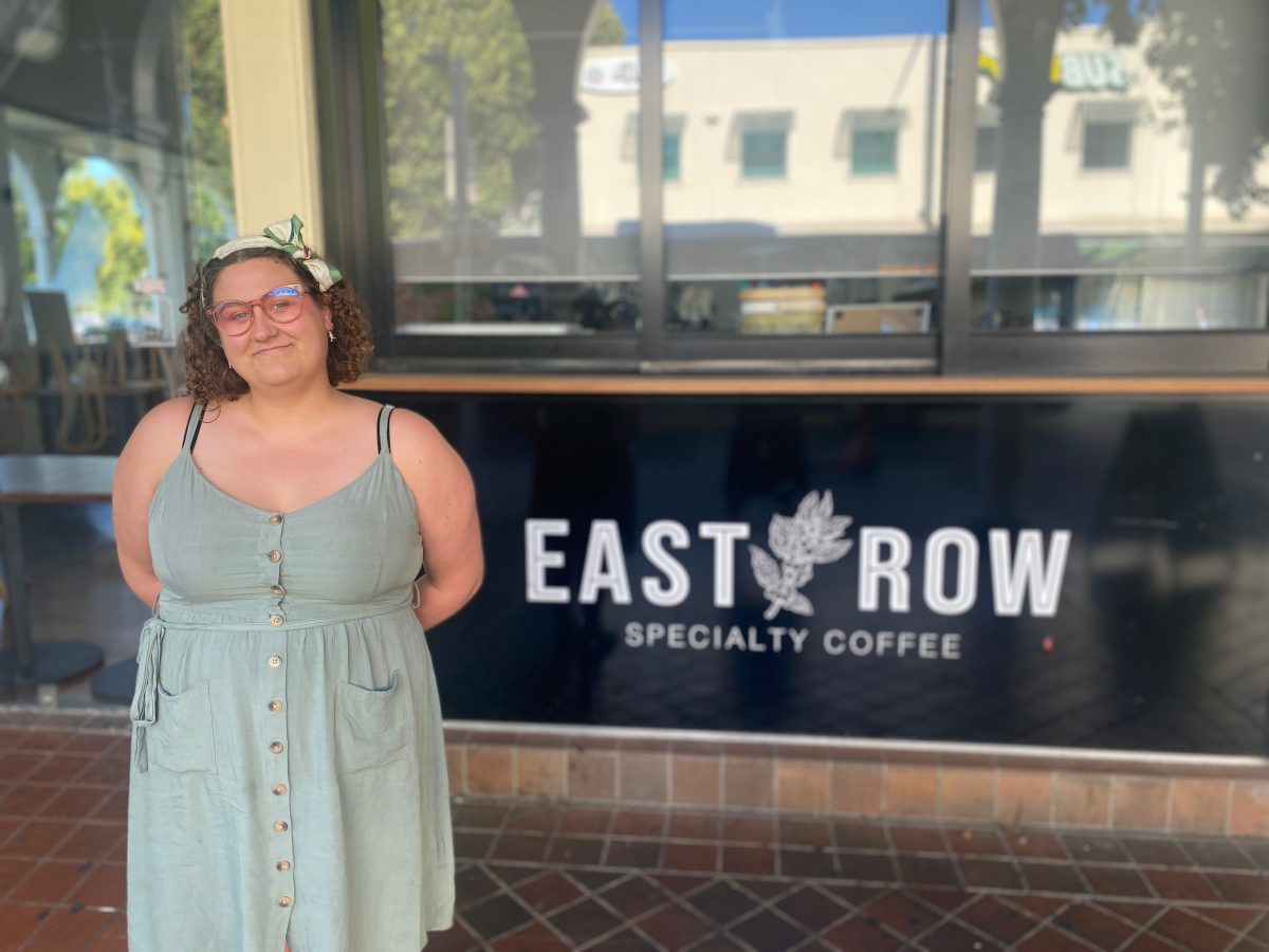 Emily wears a green dress and stands in front of the East Row logo