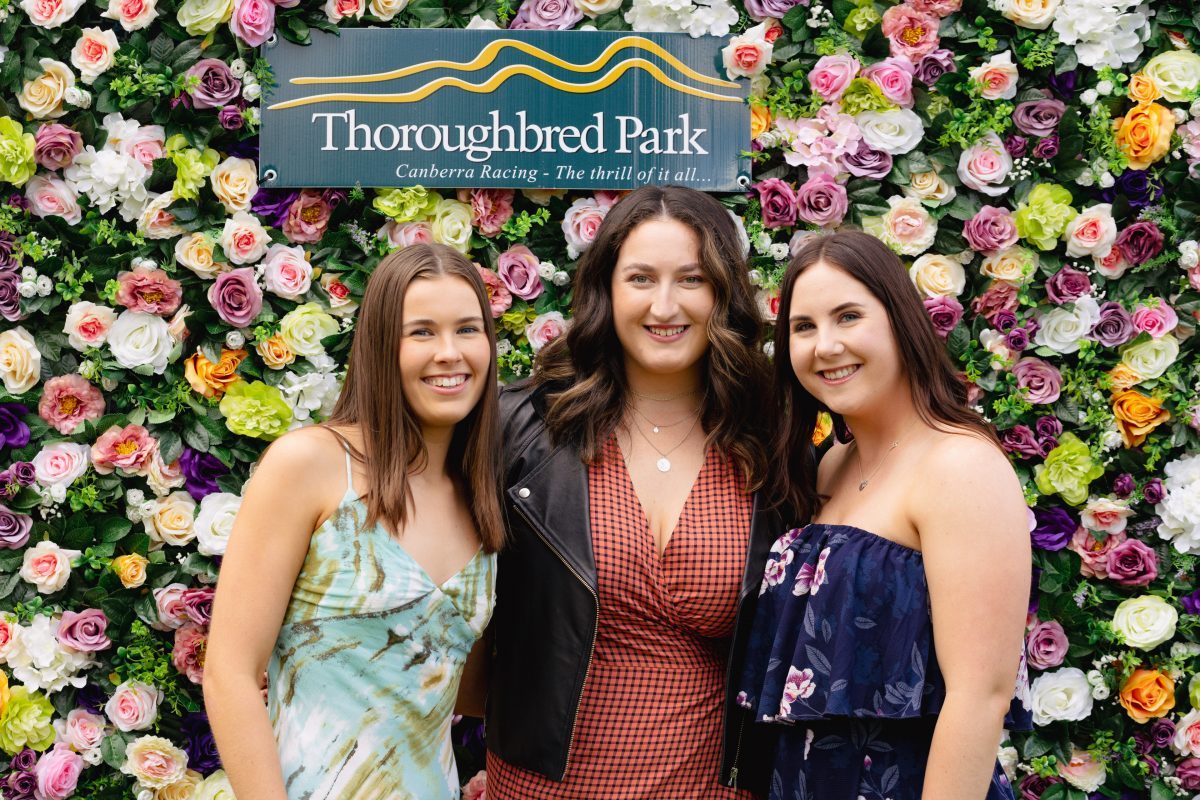 Three women standing in front of a wall of roses with a Thoroughbred Park sign hanging above their heads