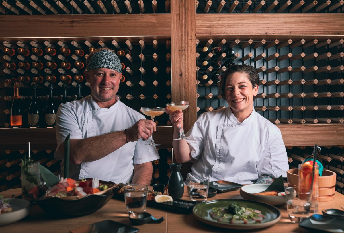 Two chefs clink glasses at table covered in food