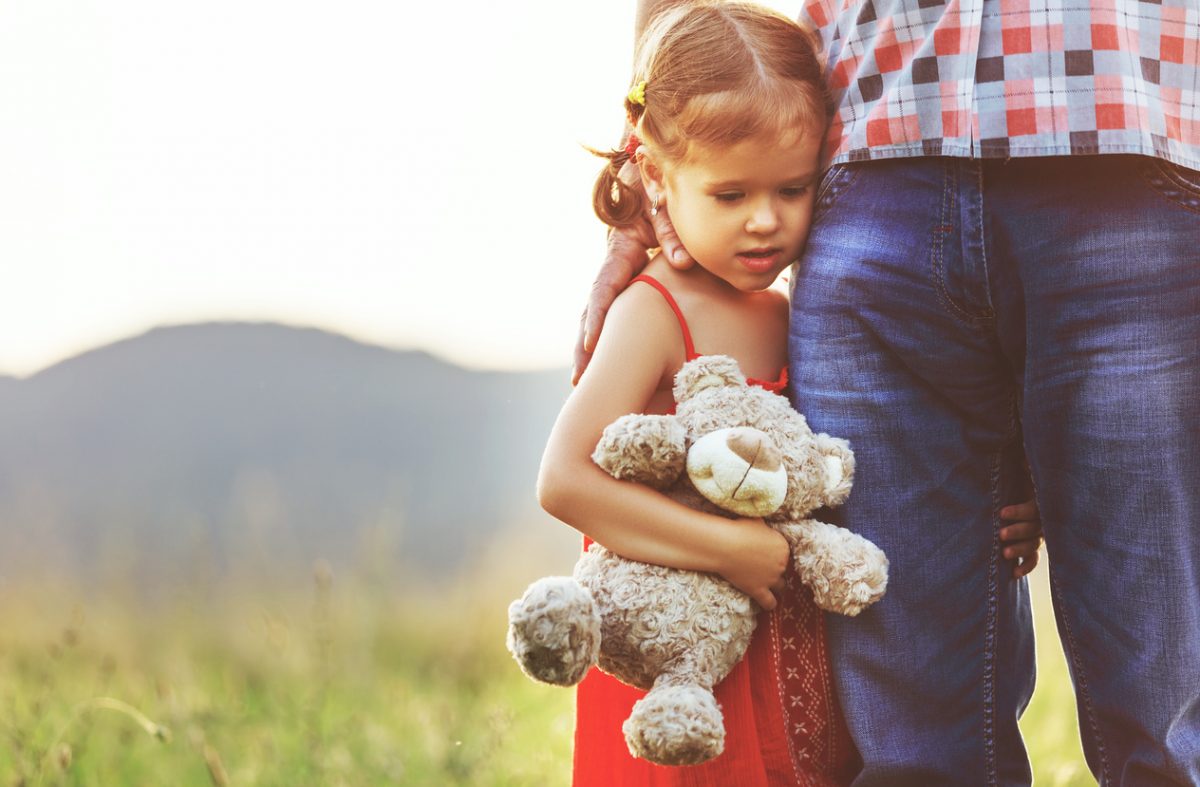 A child with a teddy hugs her father's leg