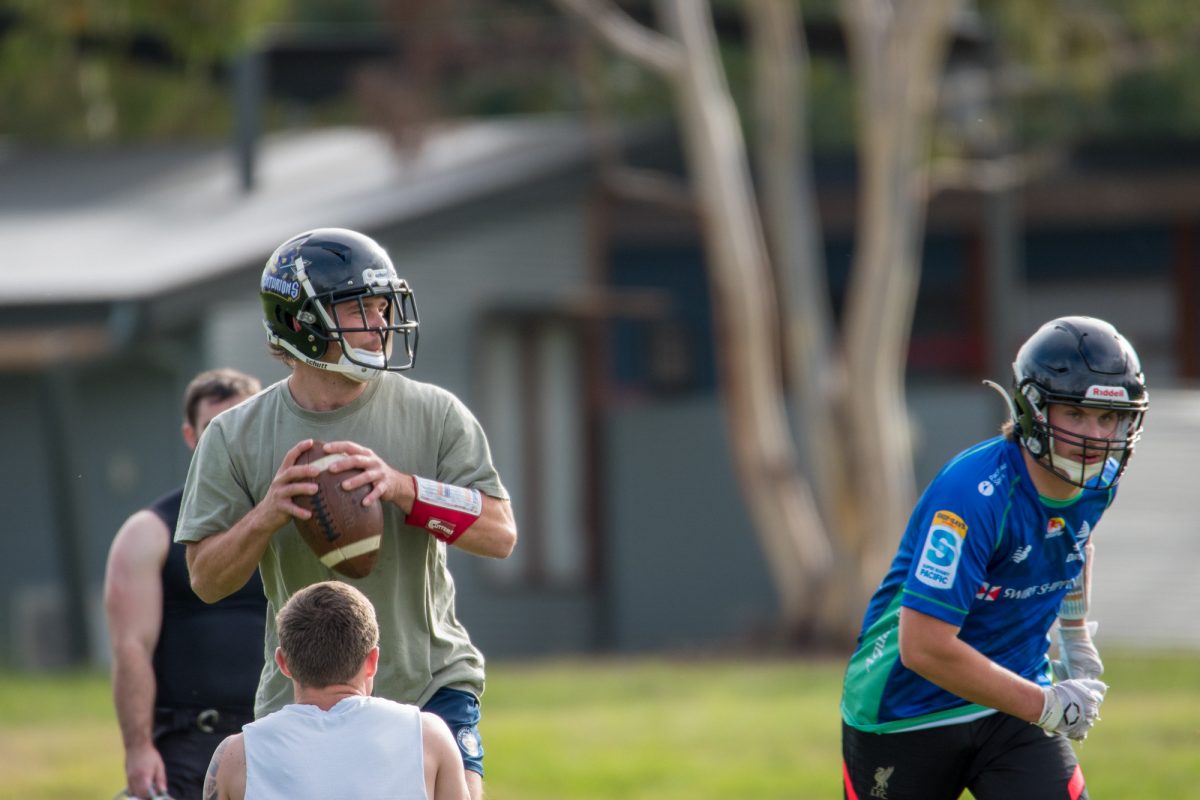 Irvin holding a ball in preparation to throw while Noble prepares to run on the right of the photo. Both are wearing their training gear with helmets with two players nearby, one in the foreground and one in the background. 