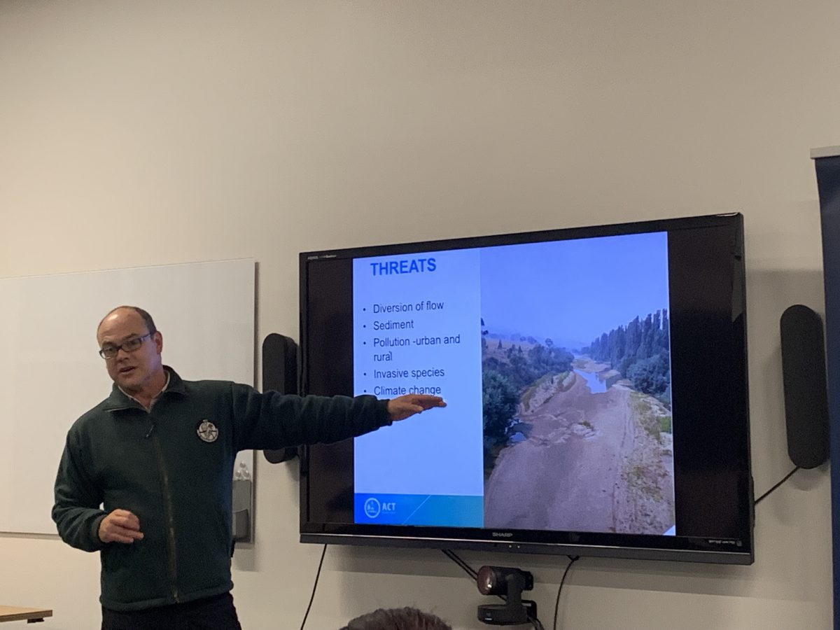 Matt Beitzel stands at the front of the room presenting with the TV power point behind him, pointing to an image of the dried up Tharwa section of the Murrumbidgee river in 2019.