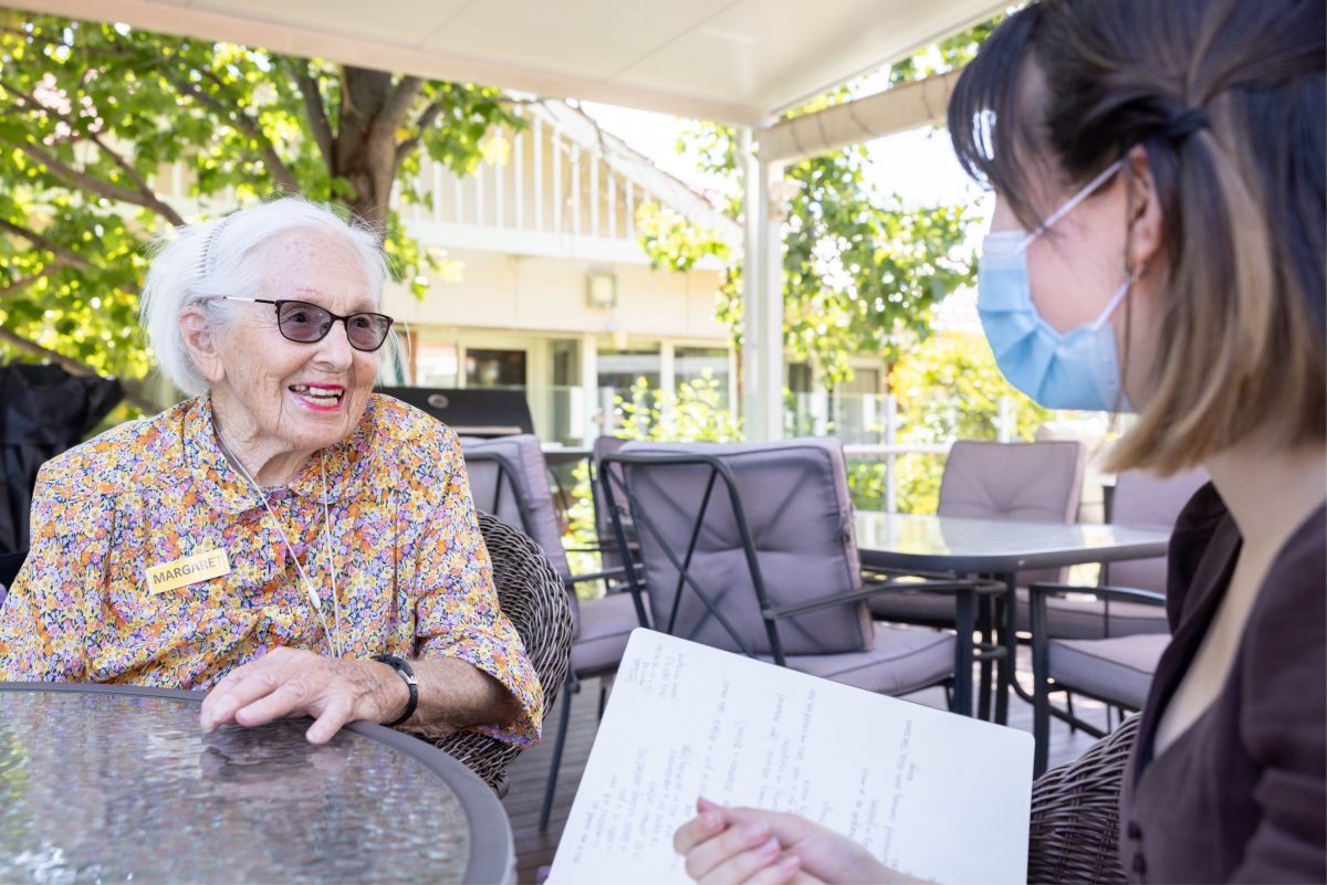 An old woman in a coloured shirt sitting opposite a young women with a blue face mask on and holding a notepad