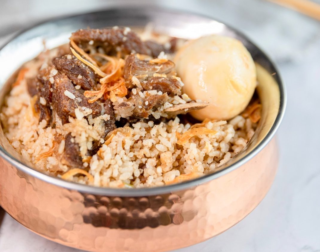 A rose tinted shiny bowl of goat biryani on a white table with cutlery in the background.