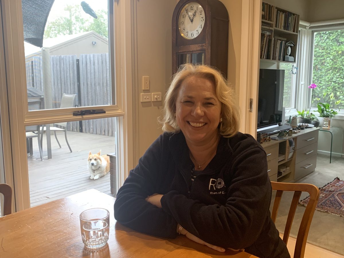 Dr. Lovett wearing a black fleece jacket sitting at the kitchen table of her home with a grandfather clock, bookcase, and window to her backyard patio behind her.
