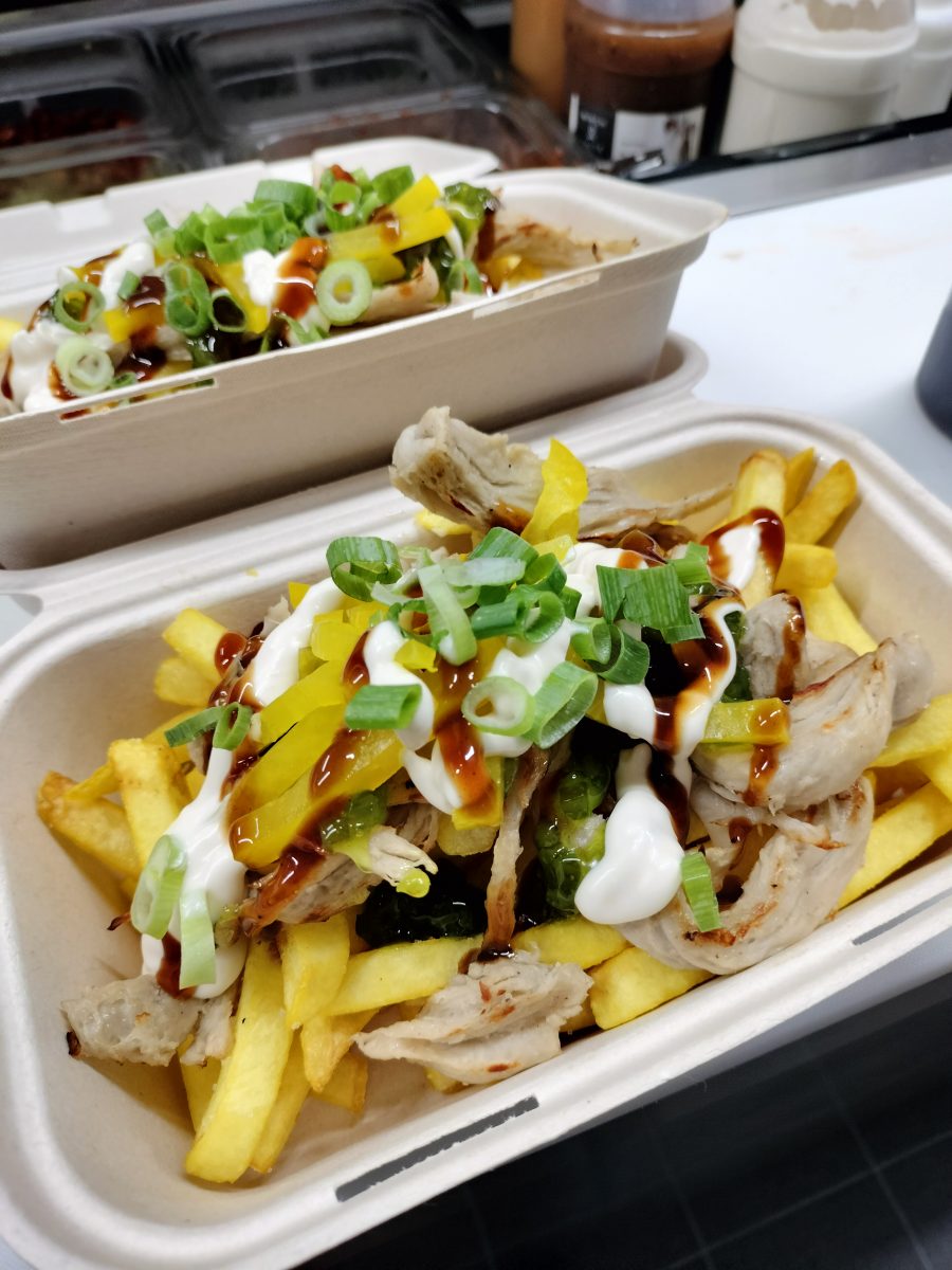 Two loaded chippies in polystyrene containers on a kitchen bench with an ingredients portion bay behind. 