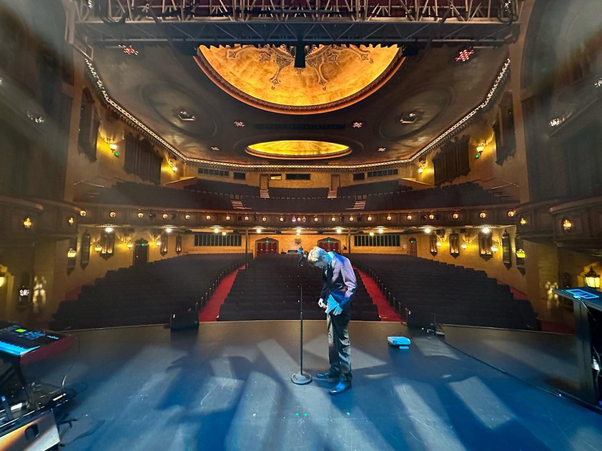 Sammy J standing centre stage pensively looking down with the microphone in front of him and in the background the empty Newcastle Civic Theatre.