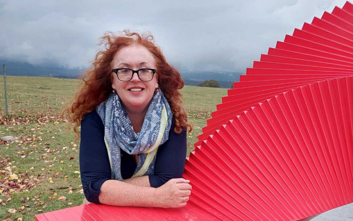 Under cloudy skies, Heidi Pritchard sits near red artwork in a field.