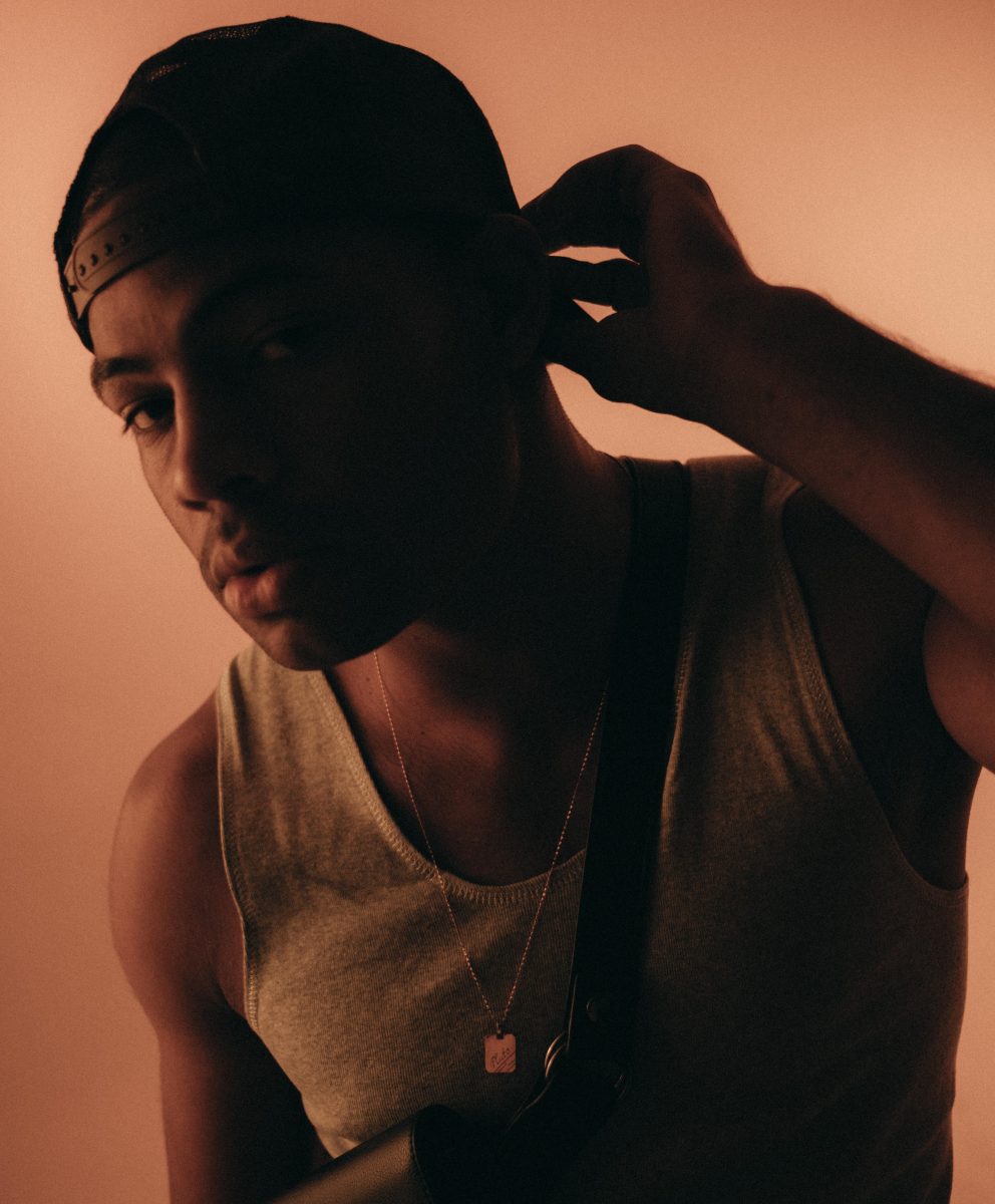 Profile shot of Ikenna wearing a white singlet, small golden chain, black satchel, and baseball cap backwards, with a bronze background and light shining onto him from the left hand side of the image.